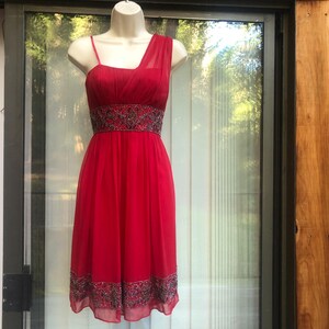 K&ouml;nnte beinhalten: Ein rotes One-Shoulder-Kleid mit einem bestickten Bund und Saum. Das Kleid wird auf einer Schaufensterpuppe pr&auml;sentiert. Das Kleid hat einen flie&szlig;enden Rock und ein eng anliegendes Oberteil. Der Hintergrund ist ein Fenster mit nat&uuml;rlichem Licht.