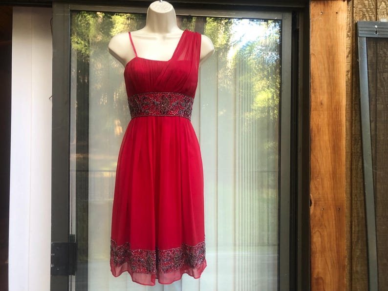 K&ouml;nnte beinhalten: Ein rotes One-Shoulder-Kleid mit einem bestickten Bund und Saum. Das Kleid wird auf einer Schaufensterpuppe pr&auml;sentiert. Das Kleid hat einen flie&szlig;enden Rock und ein eng anliegendes Oberteil. Der Hintergrund ist ein Fenster mit nat&uuml;rlichem Licht.