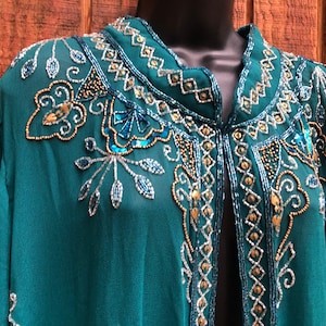 May include: Teal-colored, open-front jacket with intricate beadwork. The jacket features a mandarin collar and is embellished with silver, gold, and turquoise beads in floral and geometric patterns. The sleeves are long and flowing, and the fabric appears to be a lightweight chiffon.