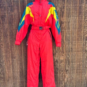 May include: A vintage red ski suit with yellow and teal accents. The one-piece suit features a high collar, a belt, and the word "FILA" on the left shoulder. The suit is hanging against a wooden background.