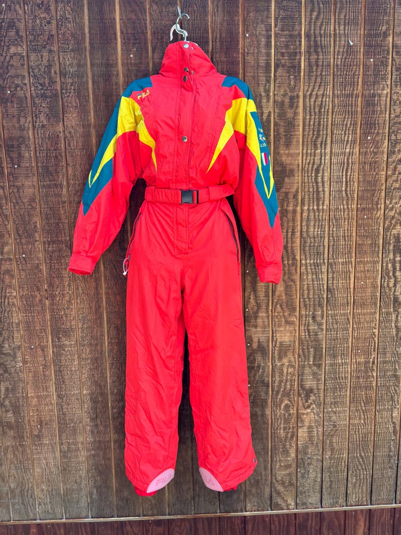 May include: A vintage red ski suit with yellow and teal accents. The one-piece suit features a high collar, a belt, and the word "FILA" on the left shoulder. The suit is hanging against a wooden background.