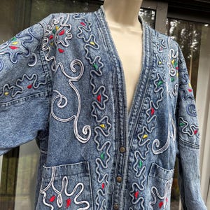 May include: A light-wash denim jacket with a V-neck design. The jacket features white, red, yellow, and green embroidered detailing. It has two front pockets and is buttoned up with bronze-colored buttons. The jacket is a vintage style.