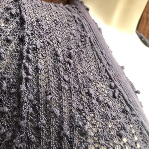 K&ouml;nnte beinhalten: Nahaufnahme eines dunkelblauen Strickkleidungsst&uuml;cks mit einem strukturierten, offen gewebten Design. Der Stoff hat ein Noppenmuster, das eine einzigartige, taktile Oberfl&auml;che erzeugt. Das Kleidungsst&uuml;ck ist &uuml;ber einer wei&szlig;en Schaufensterpuppe drapiert.