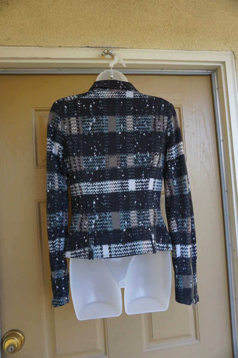 May include: A fitted jacket with a plaid pattern in black, brown, white, and teal. The jacket has a notched collar, long sleeves with zippers at the cuffs, and a tailored waist. It is displayed on a white plastic form.