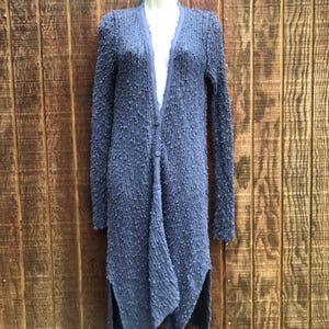K&ouml;nnte beinhalten: Ein langer, strukturierter, blauer Cardigan mit Knopfleiste und einem einzigartigen asymmetrischen Saum. Der Cardigan wird auf einer Schaufensterpuppe vor einem Holzgrund gezeigt. Die &Auml;rmel sind lang und der Stoff hat eine Noppenstruktur.