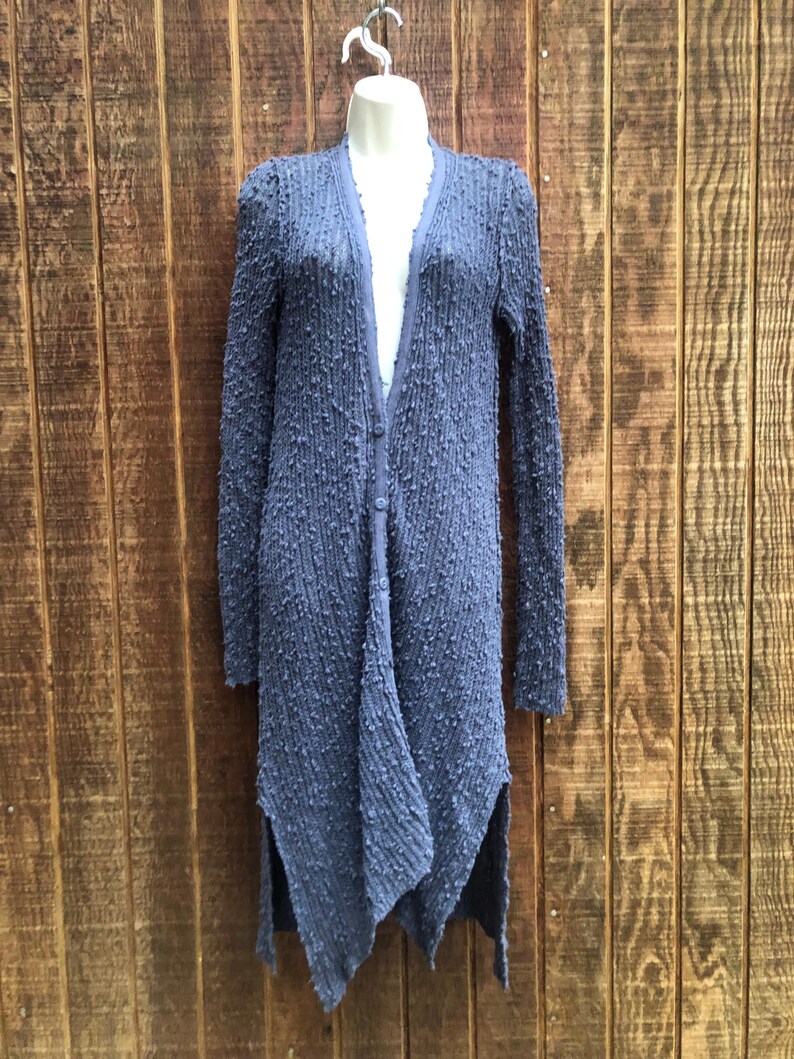 K&ouml;nnte beinhalten: Ein langer, strukturierter, blauer Cardigan mit Knopfleiste und einem einzigartigen asymmetrischen Saum. Der Cardigan wird auf einer Schaufensterpuppe vor einem Holzgrund gezeigt. Die &Auml;rmel sind lang und der Stoff hat eine Noppenstruktur.