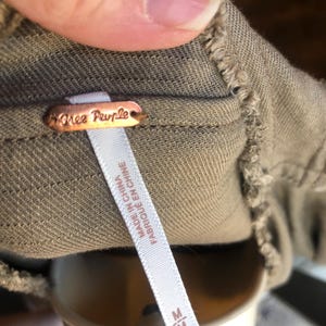 K&ouml;nnte beinhalten: Nahaufnahme eines Kleidungsst&uuml;cketiketts mit dem Markennamen "Free People" auf einem kupferfarbenen Metallanh&auml;nger. Ein wei&szlig;es Etikett darunter lautet "MADE IN CHINA FABRIQU&Eacute; EN CHINE" mit der Gr&ouml;&szlig;e "M". Der Stoff ist ein ged&auml;mpftes Olivgr&uuml;n.