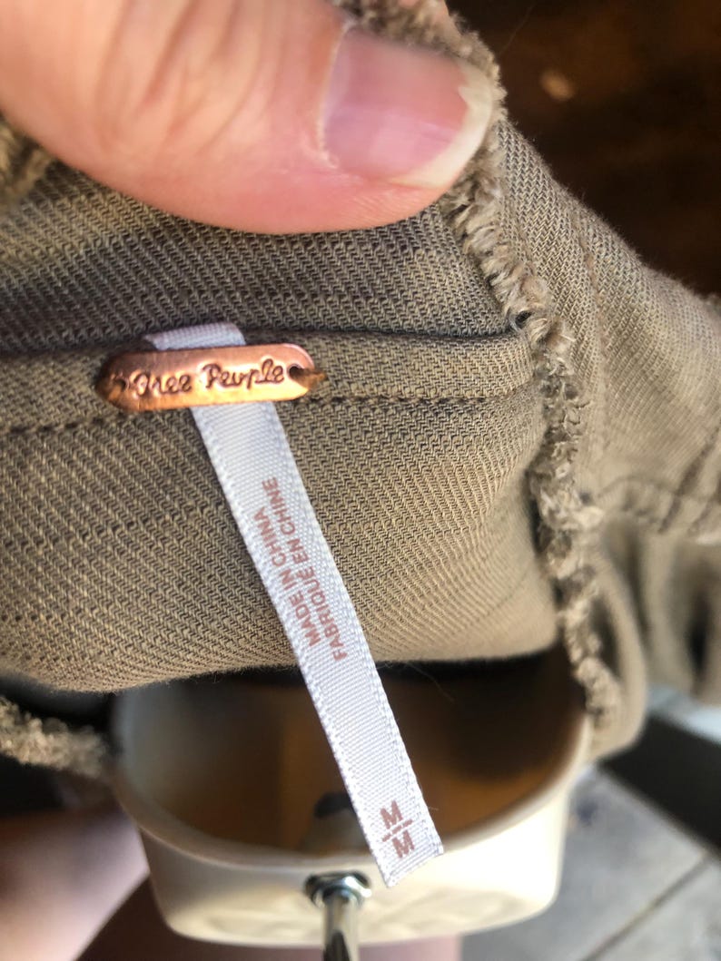 K&ouml;nnte beinhalten: Nahaufnahme eines Kleidungsst&uuml;cketiketts mit dem Markennamen "Free People" auf einem kupferfarbenen Metallanh&auml;nger. Ein wei&szlig;es Etikett darunter lautet "MADE IN CHINA FABRIQU&Eacute; EN CHINE" mit der Gr&ouml;&szlig;e "M". Der Stoff ist ein ged&auml;mpftes Olivgr&uuml;n.