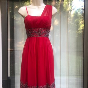 K&ouml;nnte beinhalten: Rotes One-Shoulder-Kleid auf einer Schaufensterpuppe. Das Kleid hat ein eng anliegendes Oberteil, eine mit Perlen verzierte Taille und einen flie&szlig;enden Rock. Der Saum ist ebenfalls mit Perlen verziert. Das Kleid ist aus leichtem, transparentem Stoff gefertigt.