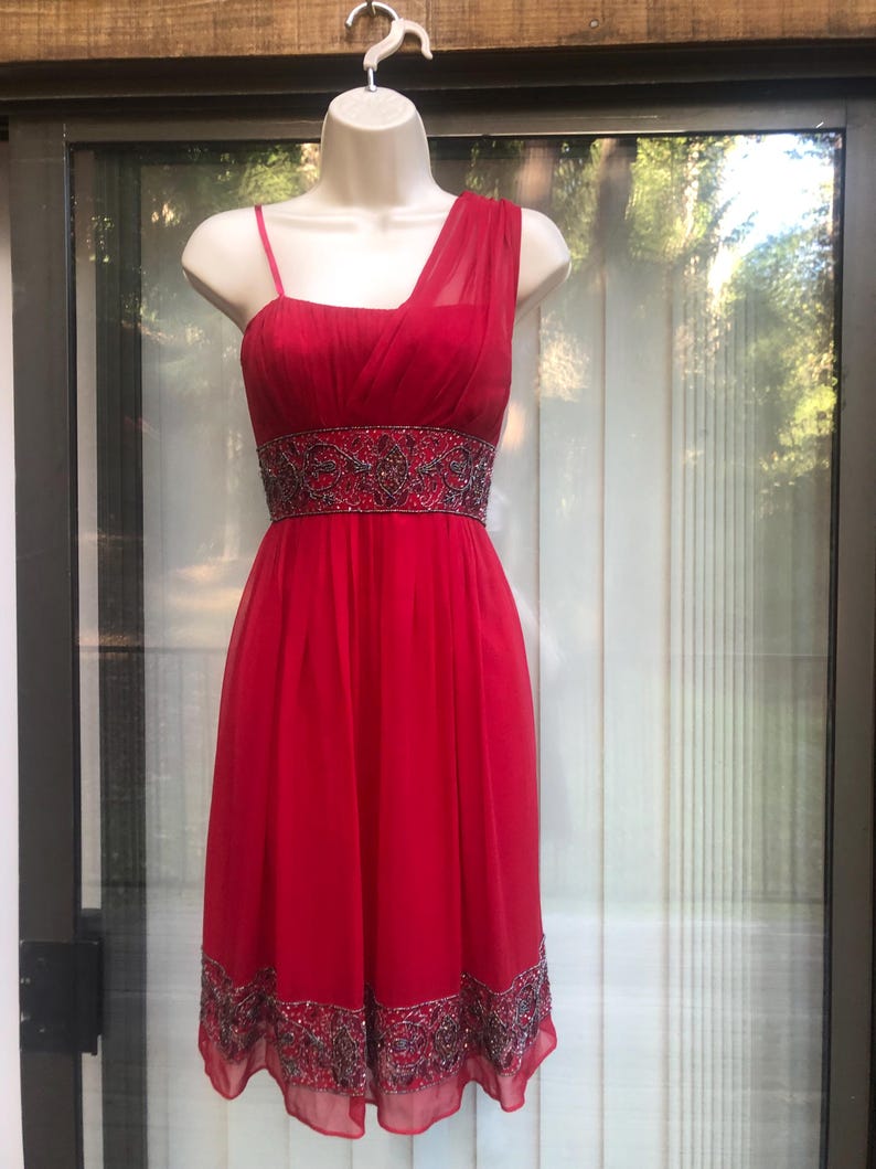 K&ouml;nnte beinhalten: Rotes One-Shoulder-Kleid auf einer Schaufensterpuppe. Das Kleid hat ein eng anliegendes Oberteil, eine mit Perlen verzierte Taille und einen flie&szlig;enden Rock. Der Saum ist ebenfalls mit Perlen verziert. Das Kleid ist aus leichtem, transparentem Stoff gefertigt.