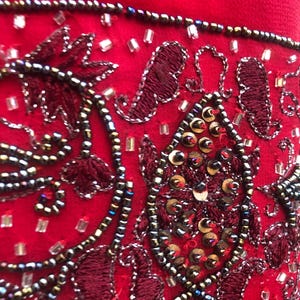 K&ouml;nnte beinhalten: Nahaufnahme eines leuchtend roten Stoffes, verziert mit aufwendigen Stickereien und Verzierungen. Das Design zeigt dunkelrote, gestickte Blumenmuster, akzentuiert mit silbernen Perlen und Pailletten. Der Gesamteffekt ist ein reichhaltiges, strukturiertes und verziertes Design.