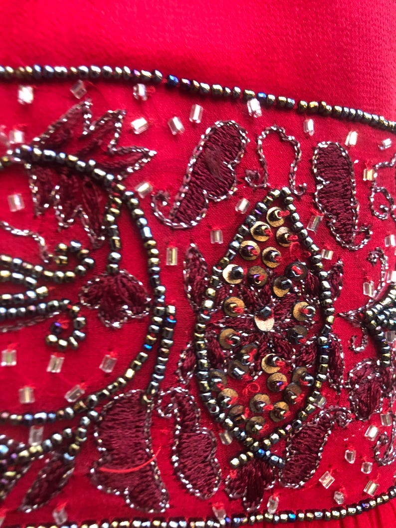 K&ouml;nnte beinhalten: Nahaufnahme eines leuchtend roten Stoffes, verziert mit aufwendigen Stickereien und Verzierungen. Das Design zeigt dunkelrote, gestickte Blumenmuster, akzentuiert mit silbernen Perlen und Pailletten. Der Gesamteffekt ist ein reichhaltiges, strukturiertes und verziertes Design.