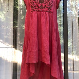 May include: A coral-colored dress with short sleeves and a high-low hemline. The dress features an embroidered floral design in brown thread around the neckline. The fabric appears to be lightweight and textured, suitable for warm weather.