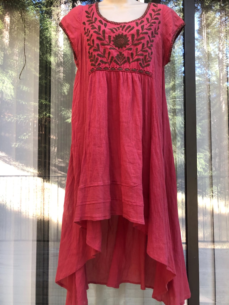 May include: A coral-colored dress with short sleeves and a high-low hemline. The dress features an embroidered floral design in brown thread around the neckline. The fabric appears to be lightweight and textured, suitable for warm weather.