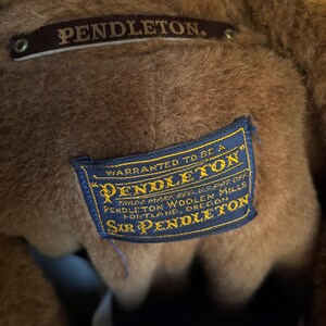 Może przedstawiać: Zbliżenie na wnętrze brązowej kurtki Pendleton. Na zdjęciu widoczne są dwie metki: brązowa metka z napisem "PENDLETON" oraz niebieska metka z napisem "WARRANTED TO BE A PENDLETON" i innym tekstem. Widoczny jest materiał kurtki.