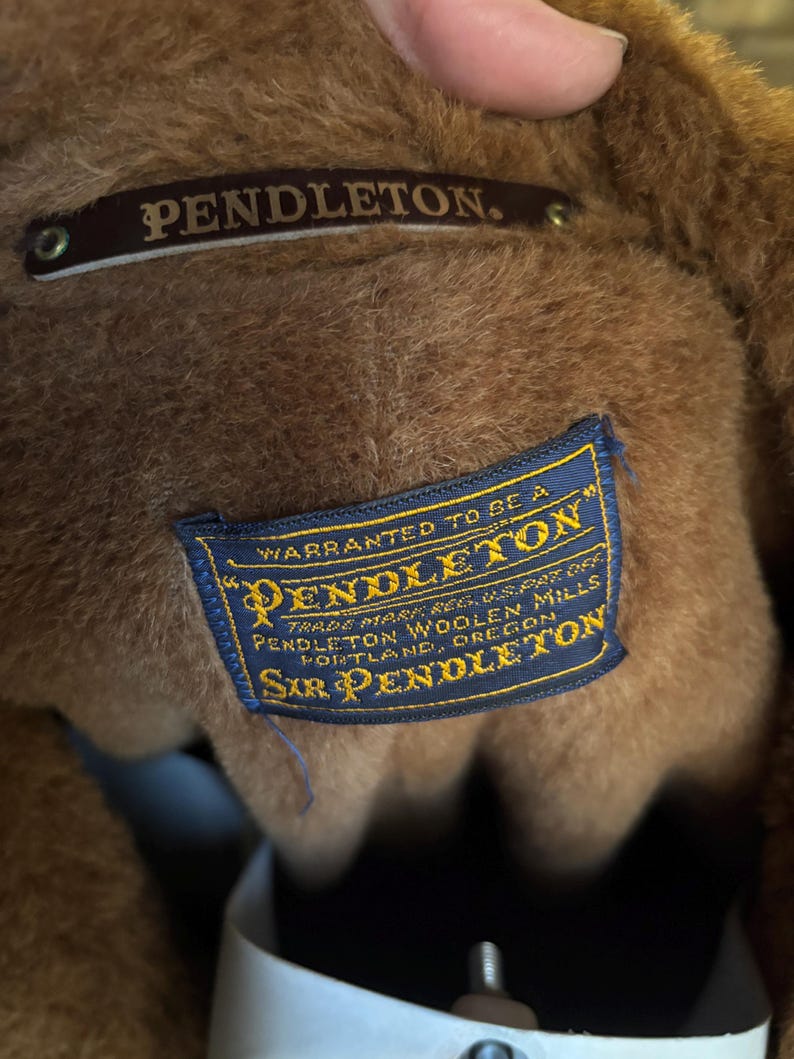 Może przedstawiać: Zbliżenie na wnętrze brązowej kurtki Pendleton. Na zdjęciu widoczne są dwie metki: brązowa metka z napisem "PENDLETON" oraz niebieska metka z napisem "WARRANTED TO BE A PENDLETON" i innym tekstem. Widoczny jest materiał kurtki.
