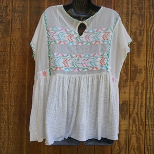 May include: White, loose-fitting tunic top with colorful embroidered geometric patterns. The top features a keyhole neckline and short sleeves with green trim. The embroidery includes pink, blue, and green accents. The tunic is displayed on a black hanger against a wooden background.