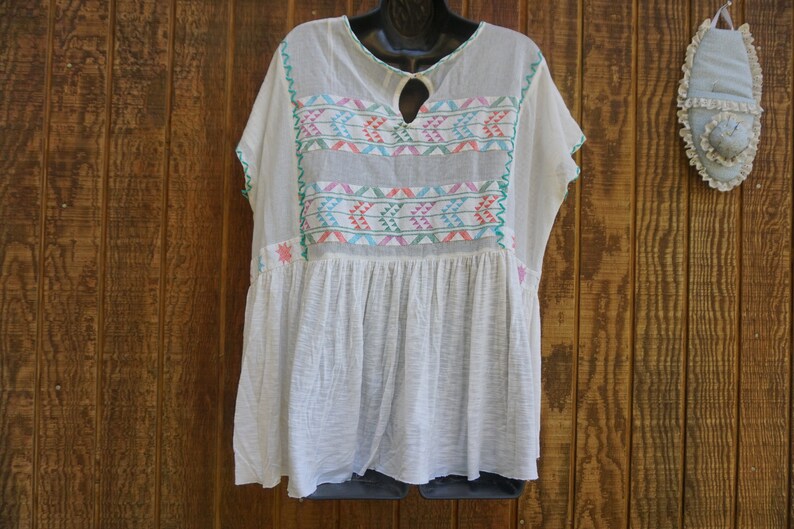 May include: White, loose-fitting tunic top with colorful embroidered geometric patterns. The top features a keyhole neckline and short sleeves with green trim. The embroidery includes pink, blue, and green accents. The tunic is displayed on a black hanger against a wooden background.