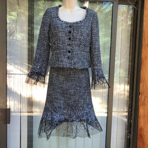 May include: A two-piece tweed suit in shades of gray and white, displayed on a mannequin. The jacket has a rounded neckline, long sleeves, and black buttons. The skirt is knee-length with a flared hem and black lace detailing.