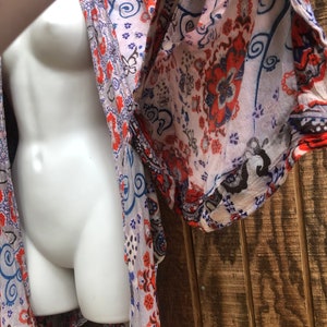 May include: A sheer, patterned kimono in shades of white, orange, blue, and black. The fabric features a floral and paisley design. The garment is draped over a white mannequin, showcasing the flowing sleeves and open front.