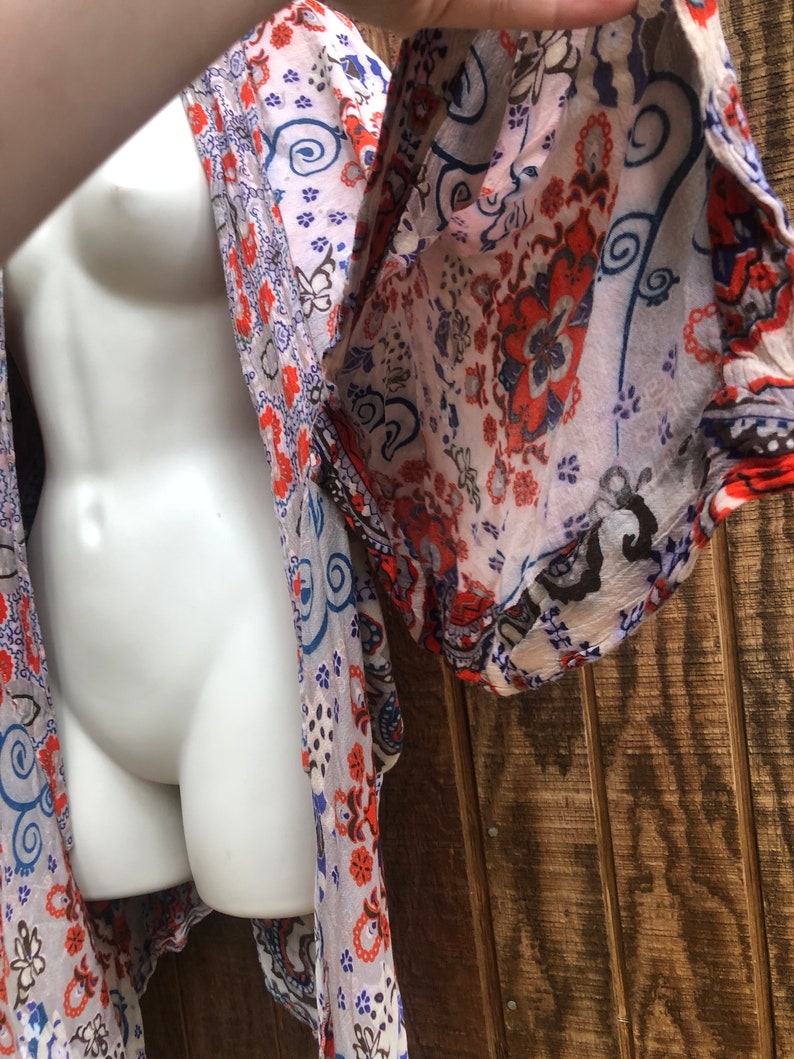 May include: A sheer, patterned kimono in shades of white, orange, blue, and black. The fabric features a floral and paisley design. The garment is draped over a white mannequin, showcasing the flowing sleeves and open front.