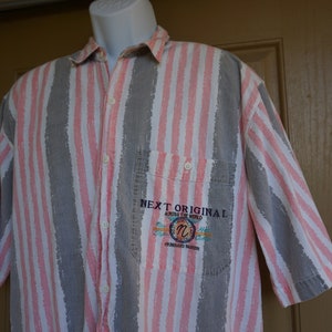 May include: Short-sleeved button-down shirt with vertical stripes in gray, white, and pink. The shirt features a chest pocket with embroidered text that reads "NEXT ORIGINAL ACROSS THE WORLD".
