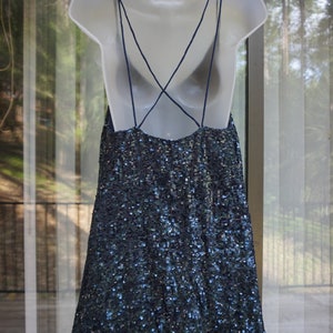 K&ouml;nnte beinhalten: Ein marineblaues Paillettenkleid mit Spaghettitr&auml;gern und tiefem R&uuml;ckenausschnitt h&auml;ngt an einer wei&szlig;en Schaufensterpuppe. Das Kleid ist mit kleinen, reflektierenden Pailletten bedeckt, die im Licht schimmern. Das Kleid hat ein schlichtes, elegantes Design.