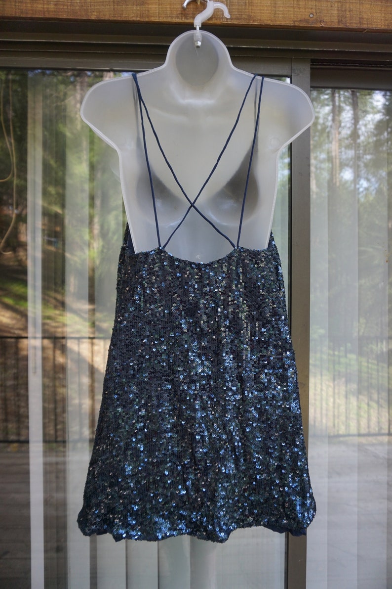 K&ouml;nnte beinhalten: Ein marineblaues Paillettenkleid mit Spaghettitr&auml;gern und tiefem R&uuml;ckenausschnitt h&auml;ngt an einer wei&szlig;en Schaufensterpuppe. Das Kleid ist mit kleinen, reflektierenden Pailletten bedeckt, die im Licht schimmern. Das Kleid hat ein schlichtes, elegantes Design.