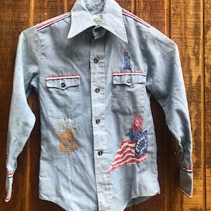 May include: A light blue long-sleeved button-down shirt with red, white, and blue trim. The shirt features embroidered images of the Statue of Liberty, the Liberty Bell, and a woman holding an American flag.