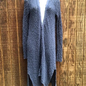 K&ouml;nnte beinhalten: Lange, strukturierte Strickjacke in ged&auml;mpftem Blau-Grau. Die Strickjacke hat lange &Auml;rmel, einen V-Ausschnitt und einen asymmetrischen Saum mit Seitenschlitzen. Der Stoff hat eine noppenartige Textur, die dem Kleidungsst&uuml;ck visuelles Interesse verleiht.