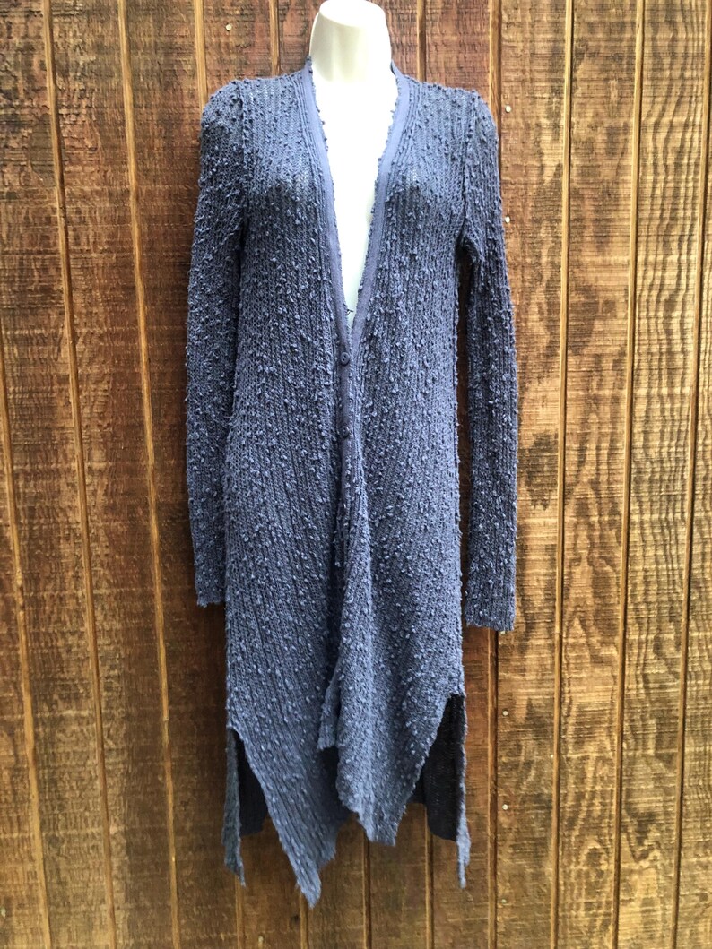 K&ouml;nnte beinhalten: Lange, strukturierte Strickjacke in ged&auml;mpftem Blau-Grau. Die Strickjacke hat lange &Auml;rmel, einen V-Ausschnitt und einen asymmetrischen Saum mit Seitenschlitzen. Der Stoff hat eine noppenartige Textur, die dem Kleidungsst&uuml;ck visuelles Interesse verleiht.