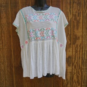 May include: White, lightweight tunic with colorful embroidered geometric patterns across the chest and sleeves. The embroidery features teal, pink, orange, and blue accents. The tunic has a loose, flowing fit and a gathered waist.