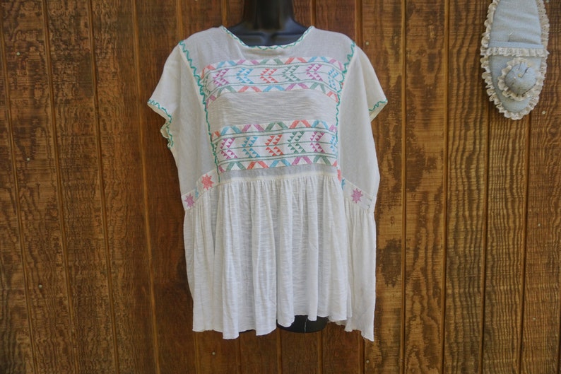 May include: White, lightweight tunic with colorful embroidered geometric patterns across the chest and sleeves. The embroidery features teal, pink, orange, and blue accents. The tunic has a loose, flowing fit and a gathered waist.