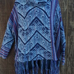 May include: A blue and white patterned poncho with a cowl neck and fringe. The poncho features a geometric design in shades of blue and white, with red accents. The bottom hem is finished with long blue fringe.
