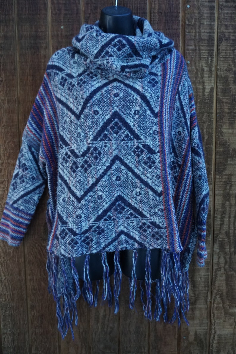 May include: A blue and white patterned poncho with a cowl neck and fringe. The poncho features a geometric design in shades of blue and white, with red accents. The bottom hem is finished with long blue fringe.