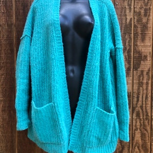May include: A teal blue open-front cardigan with long sleeves and two pockets. The cardigan is made of a soft, fuzzy material.