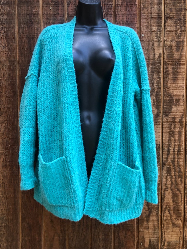 May include: A teal blue open-front cardigan with long sleeves and two pockets. The cardigan is made of a soft, fuzzy material.