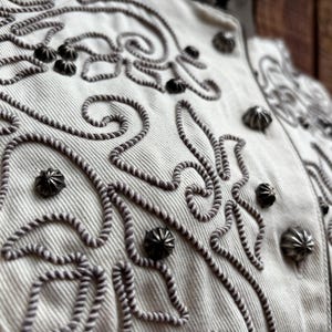 Puede incluir: Primer plano de una chaqueta color crema con intrincados bordados en forma de cuerda gris oscuro. La chaqueta presenta tachuelas met&aacute;licas decorativas y un cuello alto. El dise&ntilde;o incluye patrones florales y en espiral.