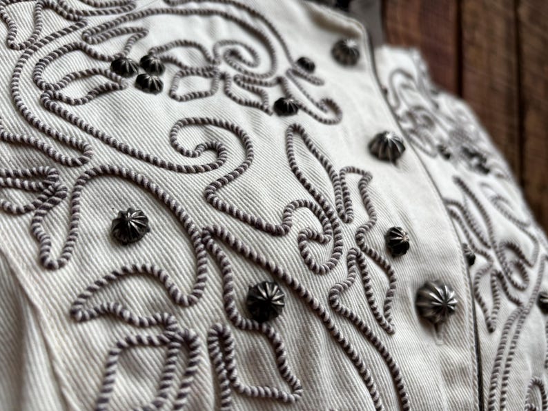 Puede incluir: Primer plano de una chaqueta color crema con intrincados bordados en forma de cuerda gris oscuro. La chaqueta presenta tachuelas met&aacute;licas decorativas y un cuello alto. El dise&ntilde;o incluye patrones florales y en espiral.