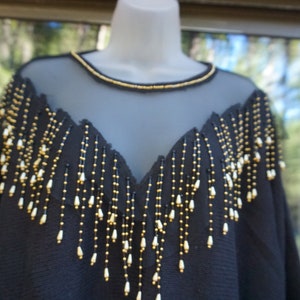 Pode incluir: Top preto de malha com franjas de contas douradas e brancas. As franjas pendem do decote e dos ombros.