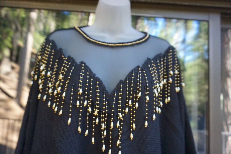 Pode incluir: Top preto de malha com franjas de contas douradas e brancas. As franjas pendem do decote e dos ombros.