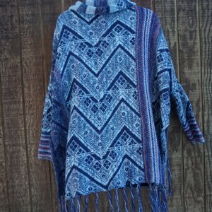 May include: A blue and white patterned poncho with a chevron design. The poncho has a fringed hem and is hanging on a black hanger against a wooden background. The pattern includes geometric shapes and zigzags.