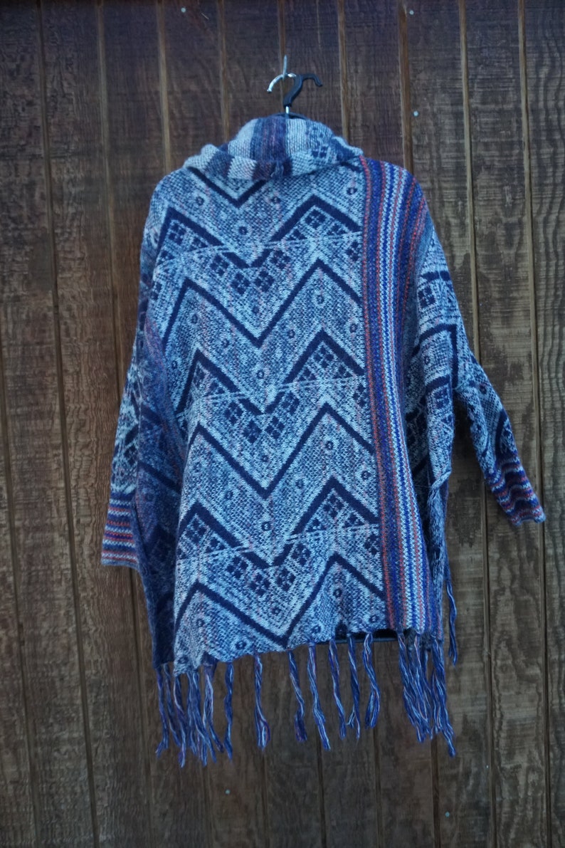 May include: A blue and white patterned poncho with a chevron design. The poncho has a fringed hem and is hanging on a black hanger against a wooden background. The pattern includes geometric shapes and zigzags.