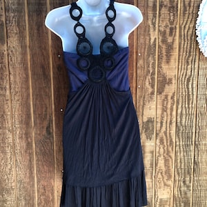 Op de afbeelding: Een marineblauwe jurk met een halterhalslijn en een zwart, cirkelvormig gehaakt ontwerp. De jurk heeft een geplooid lijfje en een gefranjerde zoom. De jurk wordt getoond op een witte mannequin tegen een houten achtergrond.