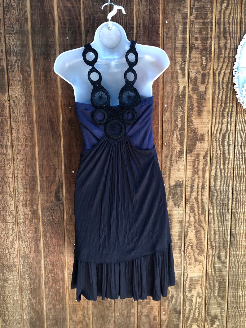 Op de afbeelding: Een marineblauwe jurk met een halterhalslijn en een zwart, cirkelvormig gehaakt ontwerp. De jurk heeft een geplooid lijfje en een gefranjerde zoom. De jurk wordt getoond op een witte mannequin tegen een houten achtergrond.