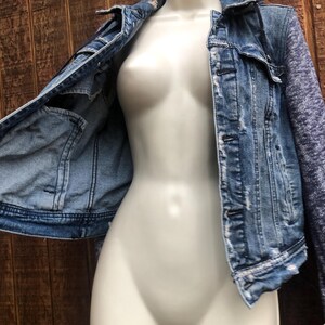 May include: A light blue denim jacket with distressed details and a contrasting heathered gray knit sleeve. The jacket is displayed on a mannequin against a wooden background. Features include front pockets and button closures.