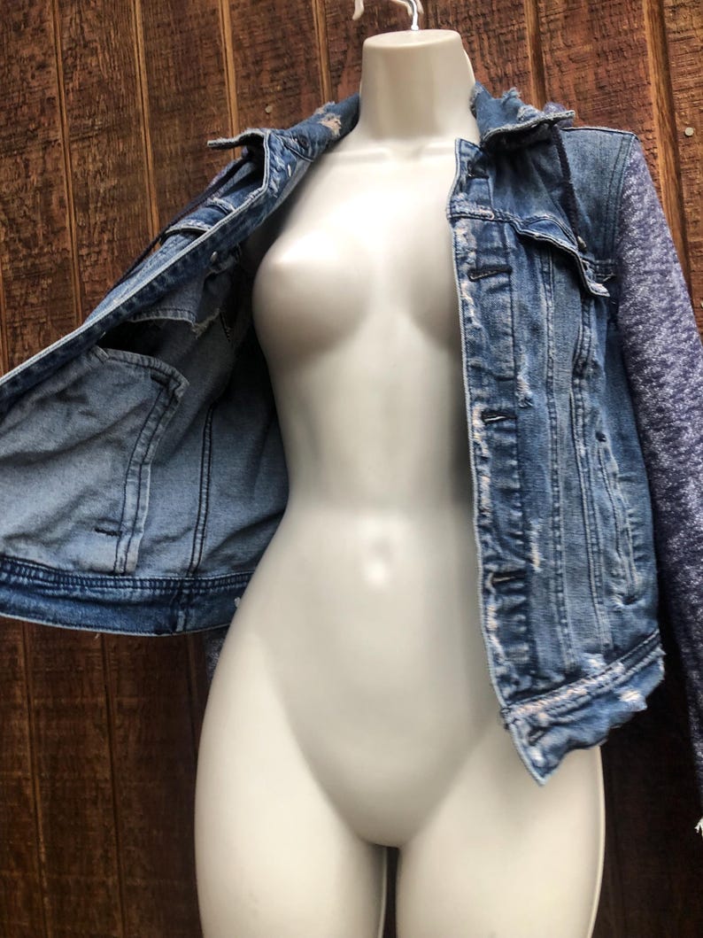 May include: A light blue denim jacket with distressed details and a contrasting heathered gray knit sleeve. The jacket is displayed on a mannequin against a wooden background. Features include front pockets and button closures.