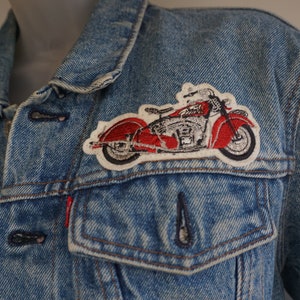 May include: A light blue denim jacket with a red and white motorcycle patch on the chest. The jacket has a classic design with button closures, a chest pocket, and a collar.