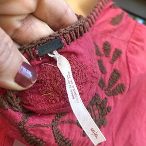May include: Close-up of a pink embroidered top with brown stitching and a brown trim. The tag reads "Free People" and "Made in India". The top has a small size tag.