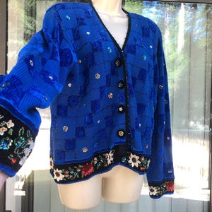 May include: A vibrant blue cardigan with a V-neck and button closure. The sweater features a checkered pattern and is embellished with sequins. The cuffs and hem are adorned with floral embroidery and black trim. The sleeves are a solid blue.